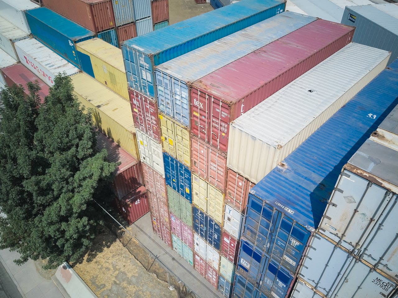 An aerial shot of multi-colored stacked shipping containers near a tree.